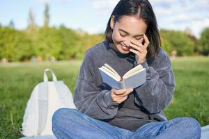 skön kvinna Sammanträde i parkera med bok. leende asiatisk flicka läsning och skrattande, avkopplande utomhus foto