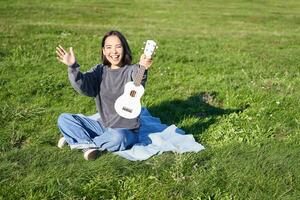 musik och instrument. glad flicka musiker, asiatisk kvinna håll henne ukulele, spelar i parkera och vågor hand på du, säger Hej till en vän foto