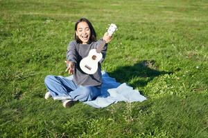 musik och instrument. porträtt av söt asiatisk flicka visar henne vit ukulele, spelar i parkera medan Sammanträde avslappnad på filt, njuter solig dag foto