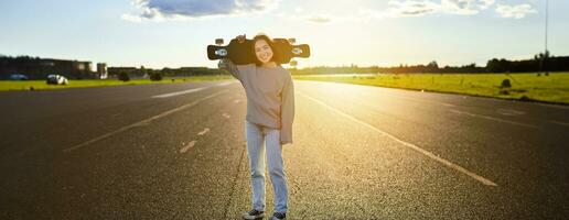 asiatisk flicka med skateboard stående på väg under solnedgång. skater Framställ med henne lång styrelse, kryssare däck under Träning foto