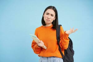 irriterad ung asiatisk kvinna, studerande komplanerar, skakningar hand och utseende besviken, står med ryggsäck och anteckningsböcker, blå bakgrund foto