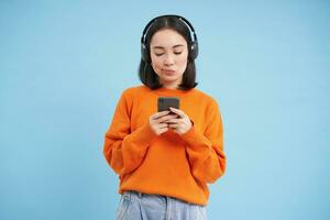 ung kvinna i hörlurar och med smartphone lyssnar musik, åtnjuter sång favorit låtar i henne Spellista, blå bakgrund foto