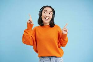 Lycklig kinesisk kvinna i hörlurar, lyssnar musik, åtnjuter favorit låt i henne Spellista, står över blå bakgrund foto