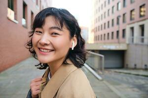 gående runt om stad. leende eleganta koreanska flicka i hörlurar, lyssnar till musik medan promenader på gata, användningar smartphone app, spela låtar foto