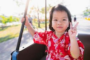 glad tjej rider i en golfbil i parken under våren. sött leende baby höjer två fingrar. bedårande barn som bär en röd japansk stil klänning. barn 1-2 år. foto