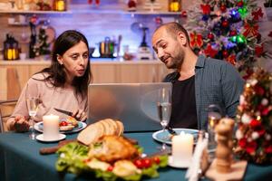 Lycklig familj äter utsökt xmas middag Sammanträde på dining tabell i x-mas dekorerad kök njuter vinter- säsong. familj umgänge med vänner på dator bläddring på social media foto