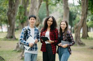 tre ung högskola studenter är läsning en bok medan avkopplande Sammanträde på gräs i en campus parkera med henne vänner. utbildning begrepp foto