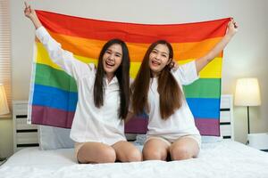 porträtt av Lycklig asiatisk lesbisk par Sammanträde med regnbåge flagga Sammanträde säng bredvid de fönster. glad två ung vuxen kvinnor levande tillsammans med relation begrepp. lgbtq livsstil stolthet månad foto