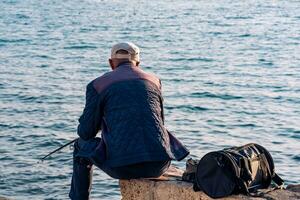 äldre fiskare sitter med en fiske stång på en kust sten foto