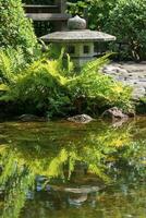 sten lykta är reflekterad i de vatten av en damm i en japansk trädgård foto
