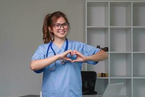 skön asiatisk kvinna bär läkare enhetlig och stetoskop leende i kärlek håller på med hjärta symbol form med händer. romantisk begrepp. foto