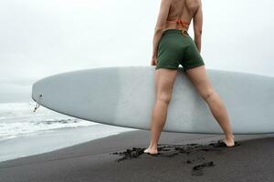 kvinna surfare innehav surfingbräda stående på svart sandig strand. låg vinkel se av ben och rumpa foto