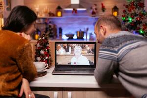 Lycklig familj talande med avlägsen mormor under uppkopplad videosamtal möte telefonkonferens Sammanträde på tabell i xmas dekorerad kök. glad par fira jul Semester foto