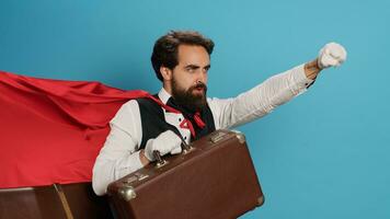 hotell piccolo verkande som övermänsklig med cape, erbjudande till hjälp människor med resväska bagage i studio. ung vuxen dörrvakt presenter komisk hjälte karaktär med röd tyg, känsla fast besluten. foto