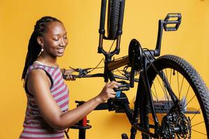 aktiva ung kvinna cyklist ser till cykel är väl förberedd för utomhus- äventyr förbi flitigt granskning de pedaler och redskap. sportig afrikansk amerikan lady gripande cykel fot spak för underhåll. foto