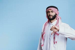 arab man bär islamic kläder, visa upp allvarlig uttryck och ger tumme ner porträtt. allvarlig muslim person som visar oenighet gest med finger och ser på kamera foto