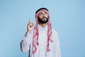 muslim man klädd i traditionell kläder pekande uppåt med finger. ung arab bär islamic thobe och rutig huvudbonad som visar och ser upp medan Framställ i studio foto