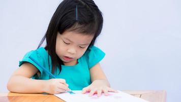 lyckligt barn med hennes imaginära konstverk. barn ritar konst och träfärgning. gullig tjej som håller blå färgpenna med höger hand. barnet ritar på papper på brunt träbord. 3-4 år gammal. foto