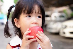 söt tjej använder handen för att hålla ett äpple och bita det med läckerhet. asiatiskt barn äter färsk röd frukt. mjukt fokus. litet barn i åldern 3 år. foto