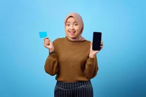 glad asiatisk kvinna som visar mobiltelefon tom skärm och håller kreditkort på blå bakgrund foto