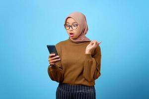 förvånad ung asiatisk kvinna tittar på mobil smartphone skärm med öppen mun på blå bakgrund