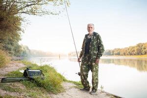 fiskare på flod porträtt. senior begrepp, pensionär, äldre man, hobby. utomhus- skön natur, fiske verktyg, fiske stång och tackla. utseende in i de kamera foto