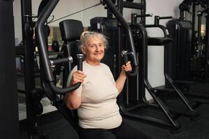 porträtt av en Lycklig pensionär i Gym. ett gammal kvinna går i för sporter på simulatorer, gläds, ler, åtnjuter, ett aktiva friska livsstil. foto