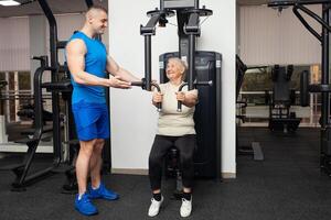 stilig ung tränare man visar senior kvinna på vilket sätt till ordentligt prestera övningar på de simulator i de Gym. Lycklig människor leende, njut av, Lycklig. aktiva sporter friska livsstil inomhus. gammal foto