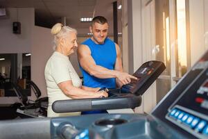 ung stilig tränare visar ett gammal kvinna på vilket sätt till klara av en löpband. Gym klasser, senior begrepp. aktiva, sportig, friska livsstil i pensionering ålder. Lycklig, leende. foto