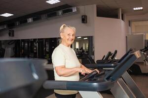 äldst kvinna av pensionering ålder kör på en löpband i de Gym. Lycklig, leende, porträtt. aktiva, atletisk, friska livsstil i gammal ålder. begrepp senior. modern teknik. foto