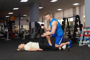 ung stilig tränare hjälper de äldre kvinna prestera stretching. kondition Gym. gymnastik. aktiva, sportig, friska livsstil i pensionering ålder. Lycklig, leende. fysisk utbildning för seniorer. liggande foto