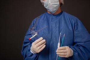 en gynekolog med en venus spegel i hans händer. gynekologisk Tillbehör, borsta, pinne. en läkare i en medicinsk mask, en morgonrock, handskar, med en gynekologisk spegel på en grå bakgrund. foto