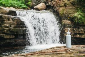vit flaska med vatten som står på en trästam mot bakgrunden av en flod och ett vattenfall. begreppet rent naturligt dricksvatten foto