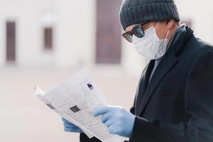 man i vinter- klädsel uppslukad i läsning en tidning, med en vit kirurgisk mask, klar indikation av pågående pandemi försiktighetsåtgärder foto