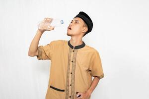 asiatisk muslim man bär muslim kläder dricka mineral vatten från en flaska. isolerat vit bakgrund. foto