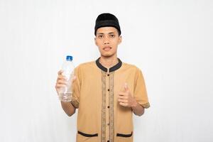 asiatisk muslim man bär muslim kläder bärande mineral vatten medan höjning hans tumme. isolerat vit bakgrund. foto