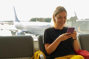 flygplats terminal. kvinna väntar för flyg använder sig av smartphone. flicka med cell telefon i flygplats surfing internet social media appar. reser kvinna i ombordstigning vardagsrum av flygbolag nav. reser flicka foto