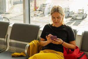 flygplats terminal. kvinna väntar för flyg använder sig av smartphone. flicka med cell telefon i flygplats surfing internet social media appar. reser kvinna i ombordstigning vardagsrum av flygbolag nav. reser flicka foto