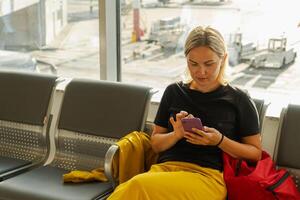 flygplats terminal. kvinna väntar för flyg använder sig av smartphone. flicka med cell telefon i flygplats surfing internet social media appar. reser kvinna i ombordstigning vardagsrum av flygbolag nav. reser flicka foto