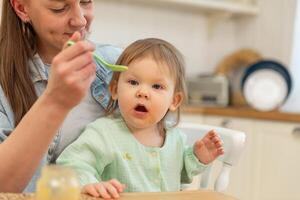 Lycklig familj på Hem. mor matning henne bebis flicka från sked i kök. liten litet barn barn med rörig rolig ansikte äter friska mat på Hem. ung kvinna mamma ger mat till unge dotter foto