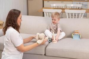 Lycklig familj på Hem. mor och bebis pojke spelar med leksaker i soffa på Hem inomhus. liten litet barn barn och barnvakt barnskötare har roligt tillsammans. ung kvinna mamma unge son resten i levande rum foto