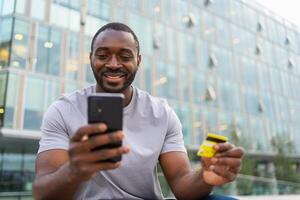 afrikansk amerikan man handla uppkopplad innehav smartphone betalande med guld kreditera kort kille på urban gata i stad uppköp på internet stiga på kreditera kort detaljer uppkopplad handla e-handel leverans service foto