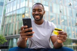 afrikansk amerikan man handla uppkopplad innehav smartphone betalande med guld kreditera kort kille på urban gata i stad uppköp på internet stiga på kreditera kort detaljer uppkopplad handla e-handel leverans service foto