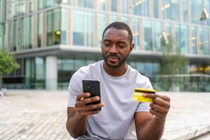 afrikansk amerikan man handla uppkopplad innehav smartphone betalande med guld kreditera kort kille på urban gata i stad uppköp på internet stiga på kreditera kort detaljer uppkopplad handla e-handel leverans service foto