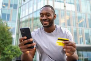 afrikansk amerikan man handla uppkopplad innehav smartphone betalande med guld kreditera kort kille på urban gata i stad uppköp på internet stiga på kreditera kort detaljer uppkopplad handla e-handel leverans service foto