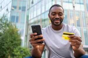 afrikansk amerikan man handla uppkopplad innehav smartphone betalande med guld kreditera kort kille på urban gata i stad uppköp på internet stiga på kreditera kort detaljer uppkopplad handla e-handel leverans service foto