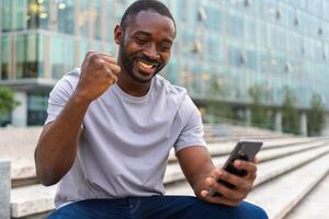 Lycklig afrikansk amerikan man euforisk vinnare med smartphone på gata i stad. person kille ser på cell telefon läsning bra Nyheter få Bra resultat vinnande uppkopplad bud känsla förvånad. vinnande gest foto