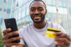 afrikansk amerikan man handla uppkopplad innehav smartphone betalande med guld kreditera kort kille på urban gata i stad uppköp på internet stiga på kreditera kort detaljer uppkopplad handla e-handel leverans service foto