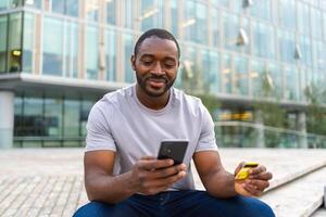 afrikansk amerikan man handla uppkopplad innehav smartphone betalande med guld kreditera kort kille på urban gata i stad uppköp på internet stiga på kreditera kort detaljer uppkopplad handla e-handel leverans service foto