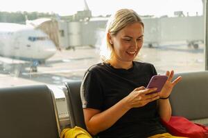 flygplats terminal. kvinna väntar för flyg använder sig av smartphone. flicka med cell telefon i flygplats surfing internet social media appar. reser kvinna i ombordstigning vardagsrum av flygbolag nav. reser flicka foto